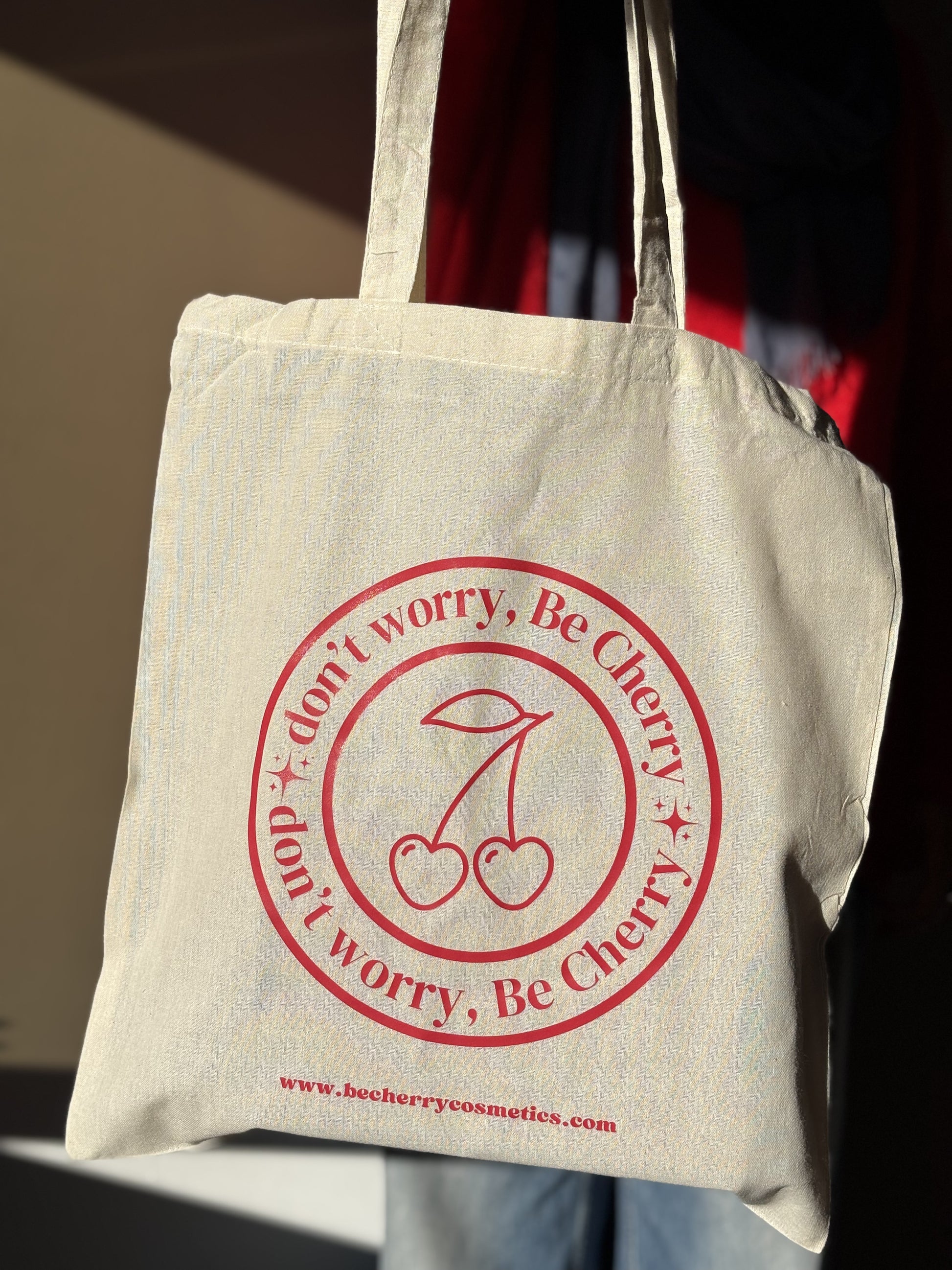 Bolsa de tela beige con asas largas, estampada con el logo "Don’t worry, Be Cherry" en rojo vibrante y dos cerezas ilustradas, ideal para llevar objetos personales, compras o libros de forma sostenible y con estilo.