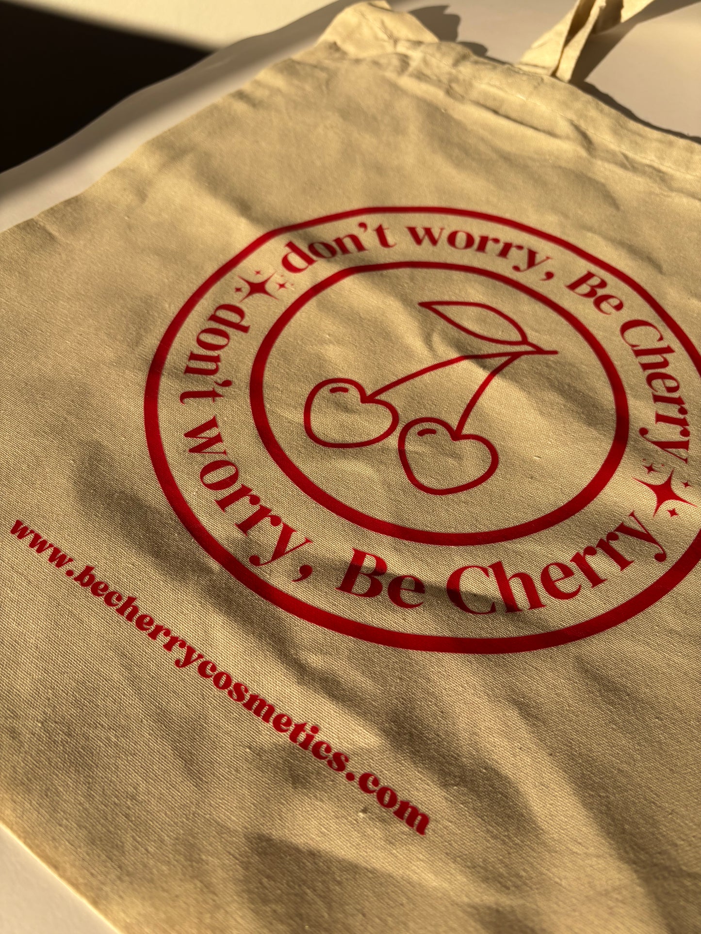 Bolsa de tela beige con asas largas, estampada con el logo "Don’t worry, Be Cherry" en rojo vibrante y dos cerezas ilustradas, ideal para llevar objetos personales, compras o libros de forma sostenible y con estilo.