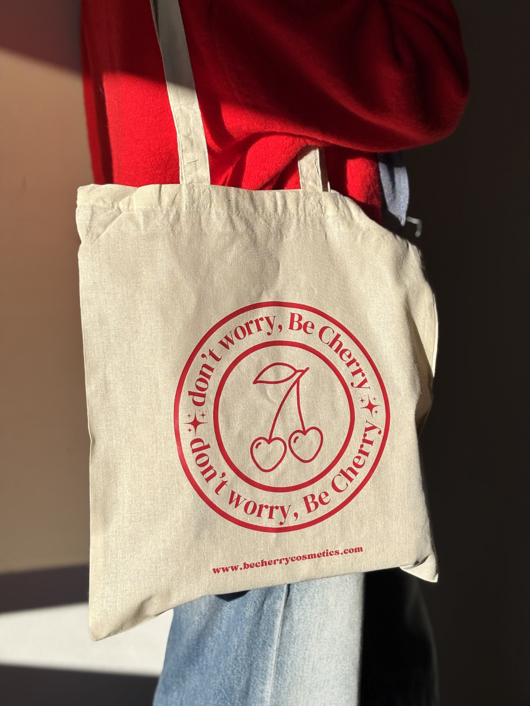 Bolsa de tela beige con asas largas, estampada con el logo "Don’t worry, Be Cherry" en rojo vibrante y dos cerezas ilustradas, ideal para llevar objetos personales, compras o libros de forma sostenible y con estilo.