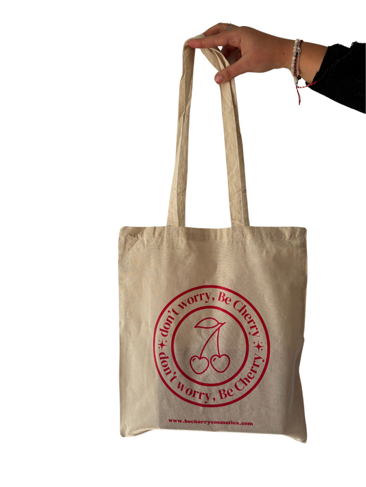 Bolsa de tela beige con asas largas, estampada con el logo "Don’t worry, Be Cherry" en rojo vibrante y dos cerezas ilustradas, ideal para llevar objetos personales, compras o libros de forma sostenible y con estilo.